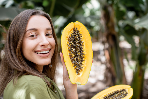 Portrait of a young woman with sliced papaya fruit on the plantation. Concept of vegetarianism, healthy eating of fresh fruits, skin care and wellbeing