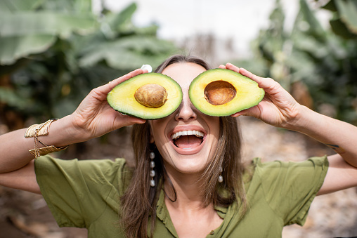 Closeup portrait of a young smiling woman hiding eyes with sliced avocado outdoors. Concept of vegetarianism, healthy eating, skin care and wellbeing