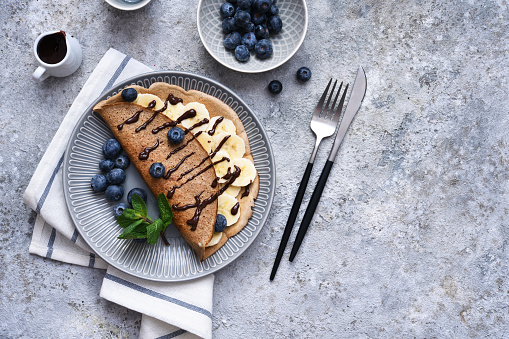 Crepe with banana, chocolate sauce and blueberries for breakfast on a stone background.