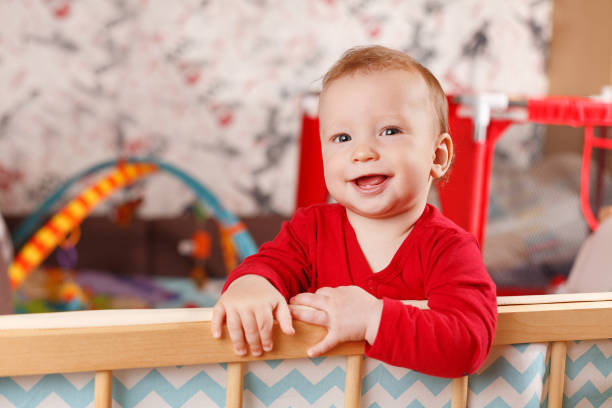 een klein kind bevindt zich en houdt de rand van het bed of de box in de kinderkamer, gelukkige en gelukkige glimlachende baby, het concept van kindontwikkeling - babybox stockfoto's en -beelden
