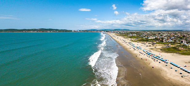 Aerial drone view of the beach, tourists, sea and waves splashing in the coast of the town Playas General Villamil, Ecuador. Sunny day. Beach houses and the horizon with some clouds in background.
