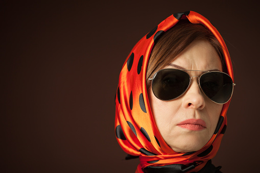 Close-up studio portrait of a 55 year old woman in an orange headdress with black polka dots and sunglasses on a brown background