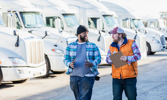 Two multi-ethnic men walking in front of a fleet of semi-trucks or tractor-trailers. The one holding the clipboard is a mature man in his 40s. The other is an African-American man in his 30s.