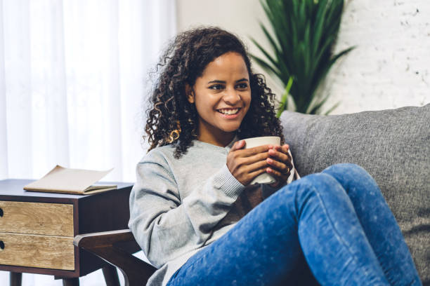 Young african american black woman relaxing drinking cup of hot coffee or tea on couch at home stock photo