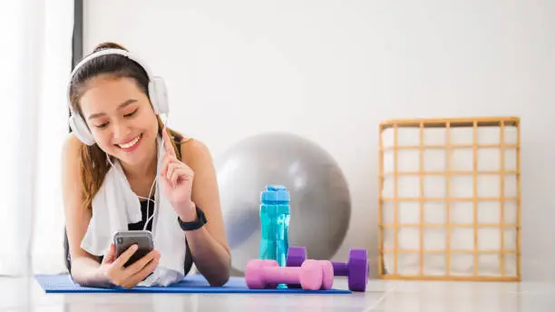 Photo of Asian woman listening to music with headphone and smartphone after play yoga and exercise at home background with copy space.Exercise for Lose weight, increase flexibility And tighten the shape.
