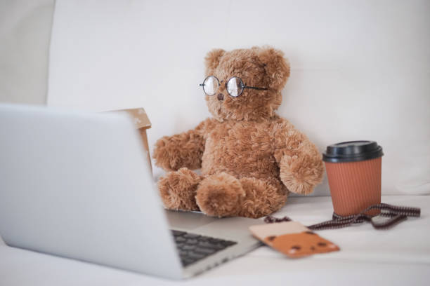 Teddy bear working concept. A cute teddy bear working in bed with a grey laptop and a cup of hot coffee stock photo
