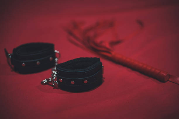 Handcuff. A black handcuff with soft fur inside and a blurred red leather whip on the background. Sex toys on the red bed sheet. soft focus. Grained and vignette filter applied. stock photo