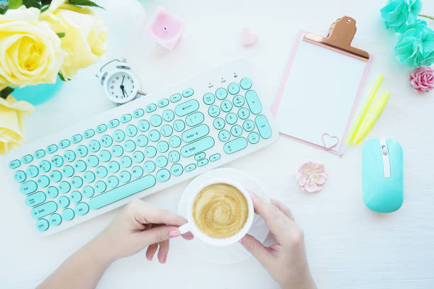 istock Female hands are holding a cup of coffee, a computer mouse and a mint-white keyboard, a pink clipboard, paper clips, a candle, an alarm clock, yellow roses. Flat layout, top view. 1206572690