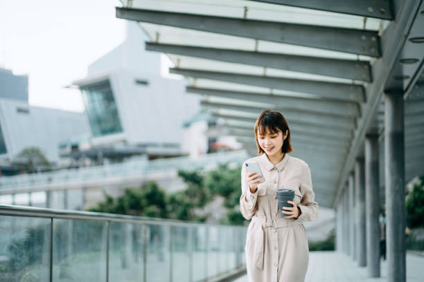 istock Young Asian woman reading text messages on smartphone and having coffee to go while walking in city bridge against urban skyscrapers 1206579168