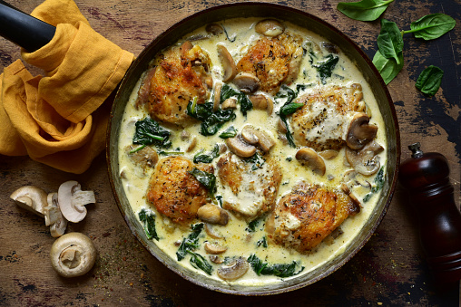 Chicken thighs stewed with mushrooms and spinach in a cream sauce in a skillet on a rustic wooden background. Top view with copy space.