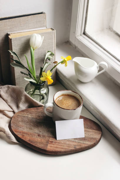 cozy easter spring still life. business card mockup scene. cup of coffee, books, wooden cutting board, milk pitcher and vase of daffodils and tulip flowers on windowsill. vertical composition. - daffodil flower yellow vase imagens e fotografias de stock