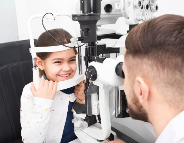 Male ophthalmologist checks the eyesight of a little girl using a binocular slit-lamp. Girl smiles because her eyesight is very good