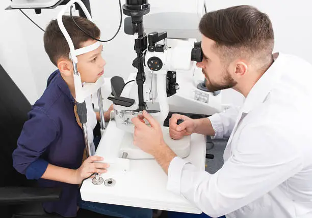 Male ophthalmologist checks eyesight of a teenage boy using a binocular slit-lamp. Checking eyesight in children