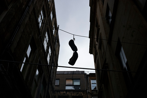 A silhouette image of shoes that are tied to each other by the shoe-strings, hangs from an electrical wire in an alley of the urban area of Glasgow, Scotland on a cloudy day.