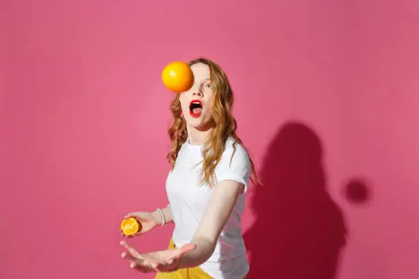 Orange fruits, healthy eating, true emotions - a young blonde woman juggling oranges on pink background - diet, people and mood concept.