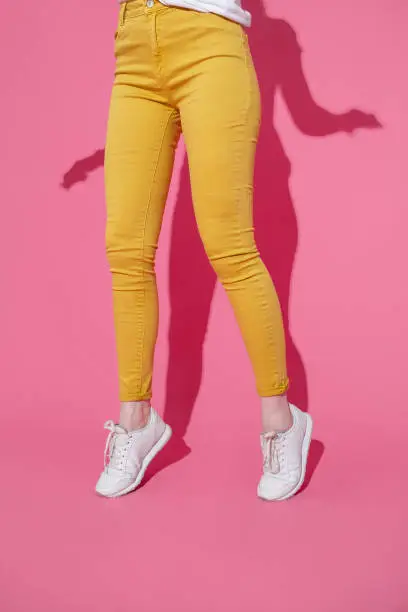 Female feet in trendy yellow jeans and white sneakers on a pink background.