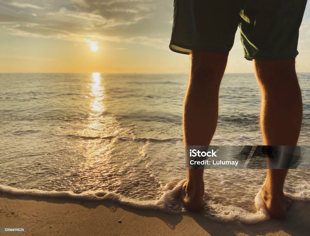 Man in shorts meets beautiful sunset, sunrise.Sea wave with foam touches legs A man in shorts meets a beautiful sunset, sunrise. A warm sea wave with foam touches the legs. Sandy beach and a serene, tranquil place on the sea. Beach Stock Photo