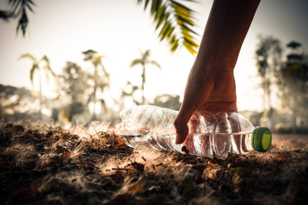 primo piano mano raccogliendo la bevanda d'acqua chiara bottiglia di plastica con un berretto verde sulla strada nel parco su sfondo sfocato, spazzatura che viene lasciata fuori dal cestino, lettiera a terra nel giardino, salva il concetto di terra - clear sky outdoors horizontal close up foto e immagini stock