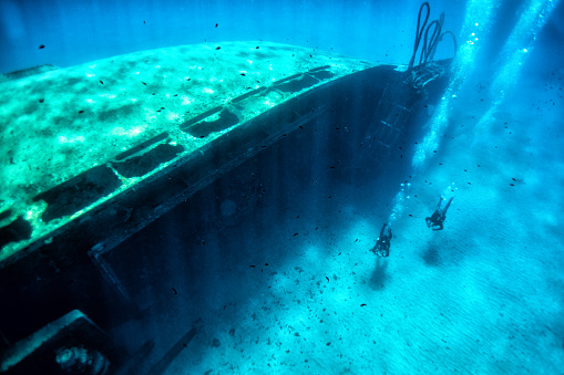 Two scuba divers exploring huge ship wreck on the sand sea bottom.
