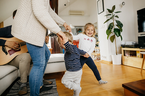 Two little kids and their mother are dancing in a living room while father is playing the guitar. They are casually dressed and it is daytime.