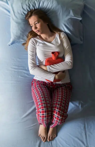 Young woman lying in pain on bed in her pajamas, cold and flu, illness, pms, menstruation, holding hot water bottle