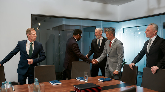 African and asian businessmen in formal wear shaking hands after signing the agreement while standing with their colleagues in the modern office. Multicultural team. Business meeting. Partnership