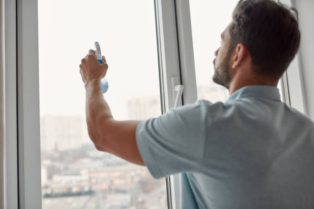 istock Professional cleaner at work. Young caucasian man in uniform cleaning window with squeegee and spray detergent while working in the living room 1206713371