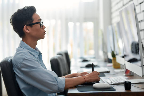istock Being creative. Side view of a young asian graphic designer in eyeglasses using digital graphics tablet while sitting at his workplace in the creative agency 1206714888