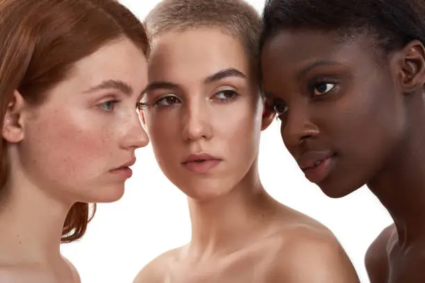 Portrait of three beautiful multicultural young women standing close to each other in studio against white background. Different ethnicity women. Different skin types. Beauty and spa