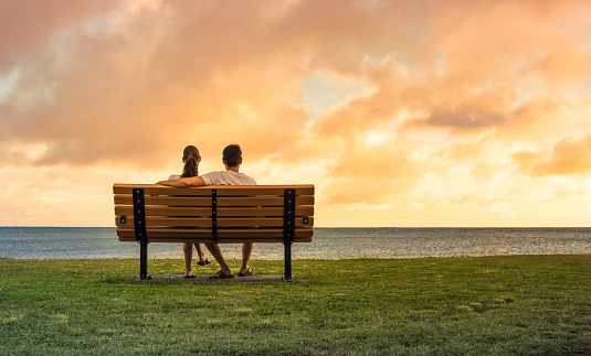 Young couple relaxing on park bench by the beach watching sunset. People love and relationships concept.