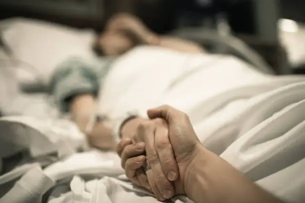 Photo of Sick woman lying in hospital bed with hand being held by love one.