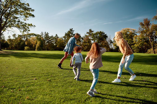 Full-length portrait of happy mother, father, little boy and girl running and playing catch game in autumn park. Family, parenthood, leisure and people concept. Horizontal shot.