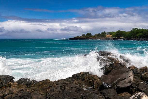 Kona Coast, Hawaii's Big Island. Wave breaking on rocky shore. Blue-green ocean. Shoreline with trees beyond. Cloudy blue sky. stock photo