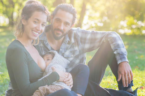A young family of mixed ethnicity are sitting together in a park on a bright summer day. Here, the mother is breastfeeding her baby boy while they sit together.
