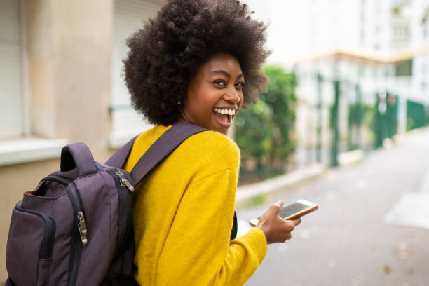 derrière de rire femme afro américaine avec sac et téléphone mobile marchant sur la rue de la ville - regarder par dessus son épaule photos et images de collection