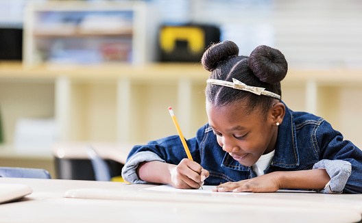 A 7 year old African-American girl in elementary school. The student is sitting at a desk in the classroom, diligently writing with a pencil on paper. She is doing schoolwork or taking a test.