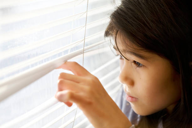 istock Little Asian girl cautiously peeking out window through blinds 1206752013