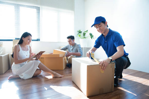 istock Delivery service worker man in blue uniform help happy couple customer packing parcel with tape in customer home.Woman with tablet signing proof document,man unpacked receive box in background. 1206764609