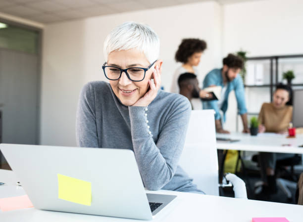 istock A senior female using her computer in an office. There are a few employees behind her at another table. 1206791568