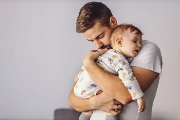 Photo of Handsome caring father hugging and holding his sleeping beloved little son. Toddler is leaning on father's shoulder, having mouth opened and dreaming.