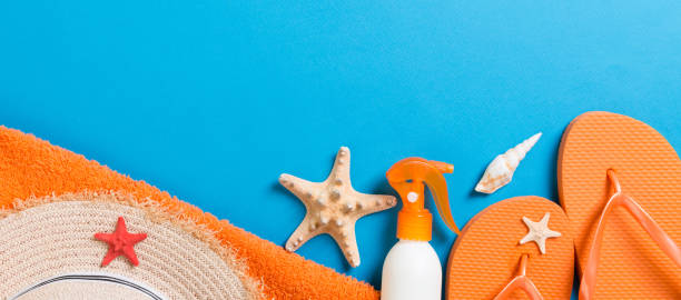 Summer beach flat lay accessories. Sunscreen bottle cream, straw hat, flip flops, towel and seashells on colored Background. Travel holiday concept with copy space