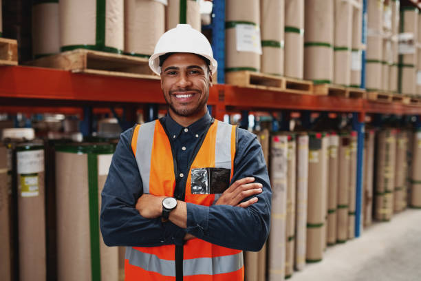 confident african american man standing with folded arms in manufacturing unit with white helmet and uniform in factory - plant stand imagens e fotografias de stock