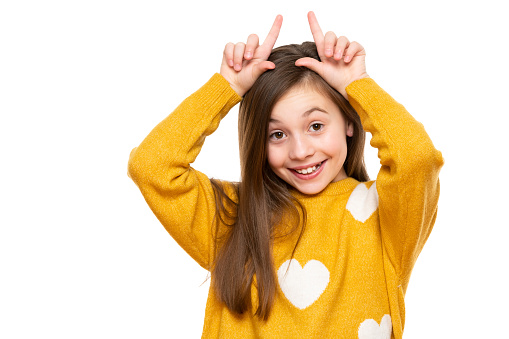 Studio shot of expressive cute child holding fingers behind head like bunny ears or horns, looking at camera and smiling. Lifestyle and beautiful people concept.