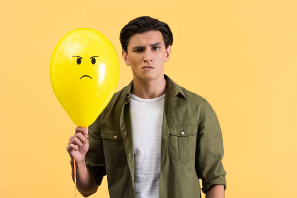 istock skeptical young man holding angry balloon, isolated on yellow 1206820491
