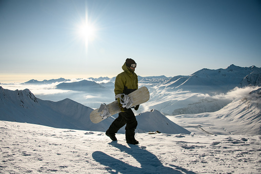 Guy with snowboard in winter clothes walks on high snowy sunny mountain top at sunny day