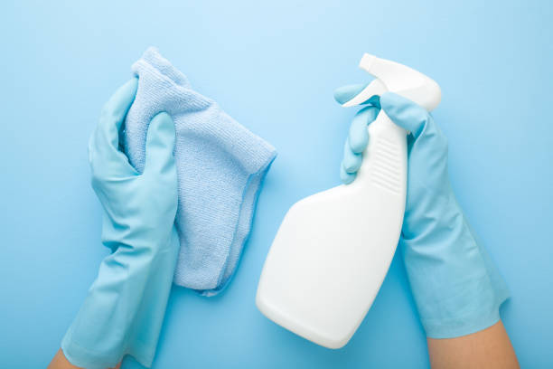 istock Hands in rubber protective gloves holding white spray bottle and rag. Detergent for different surfaces in kitchen, bathroom and other rooms. Closeup. Light pastel blue background. Point of view shot. 1206847805