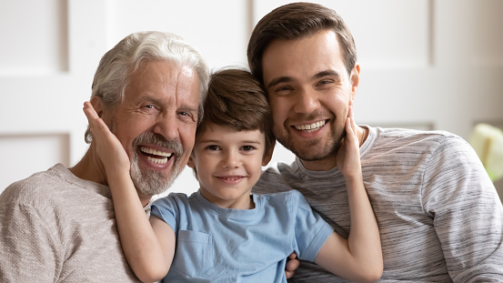 Smiling little schoolboy touching cheeks of cheerful middle aged grandfather and smiling young daddy, head shot close up portrait. Happy three generations affectionate family looking at camera.