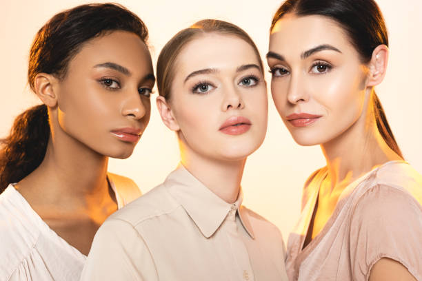 three beautiful multicultural women with makeup looking at camera isolated on beige