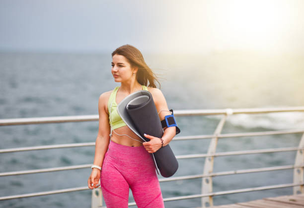 jeune femme athlétique dans le sportwear retient le tapis de yoga dans sa main et posant à l’extérieur sur la plage - aerobics beautiful bottle body photos et images de collection