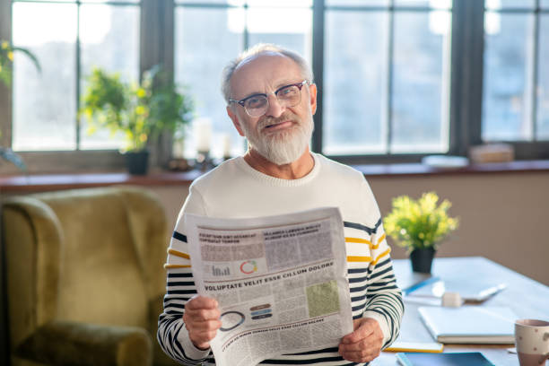 istock Grey-haired bearded man in eyewear smiling nicely while reading a newspaper 1206891376